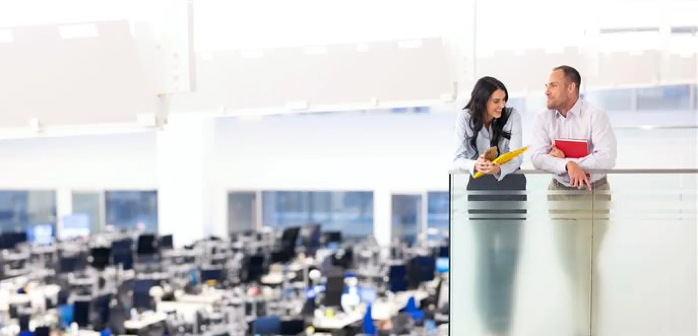 Two colleagues having a discussion on an upper floor balcony overlooking a large open-plan office filled with desks and computer monitors.