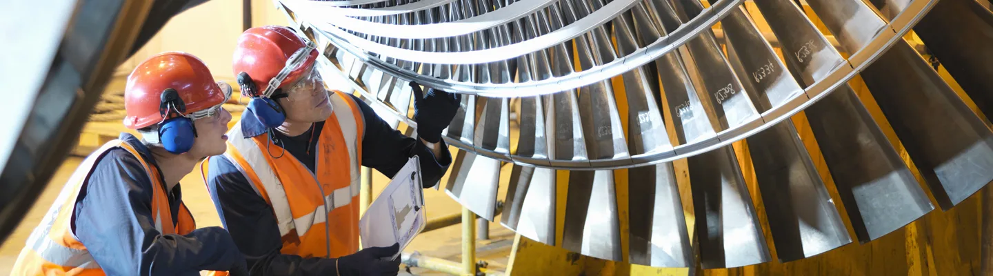 Two engineers in safety gear inspecting a large turbine blade inside an industrial facility