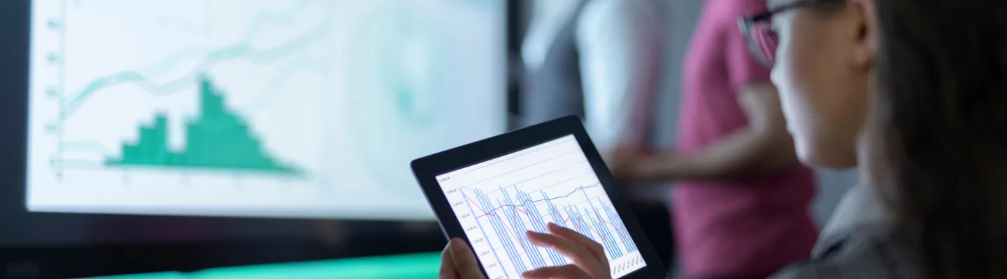A person analyzing data charts on a tablet, with a large display screen showing graphs and data visualizations in the background
