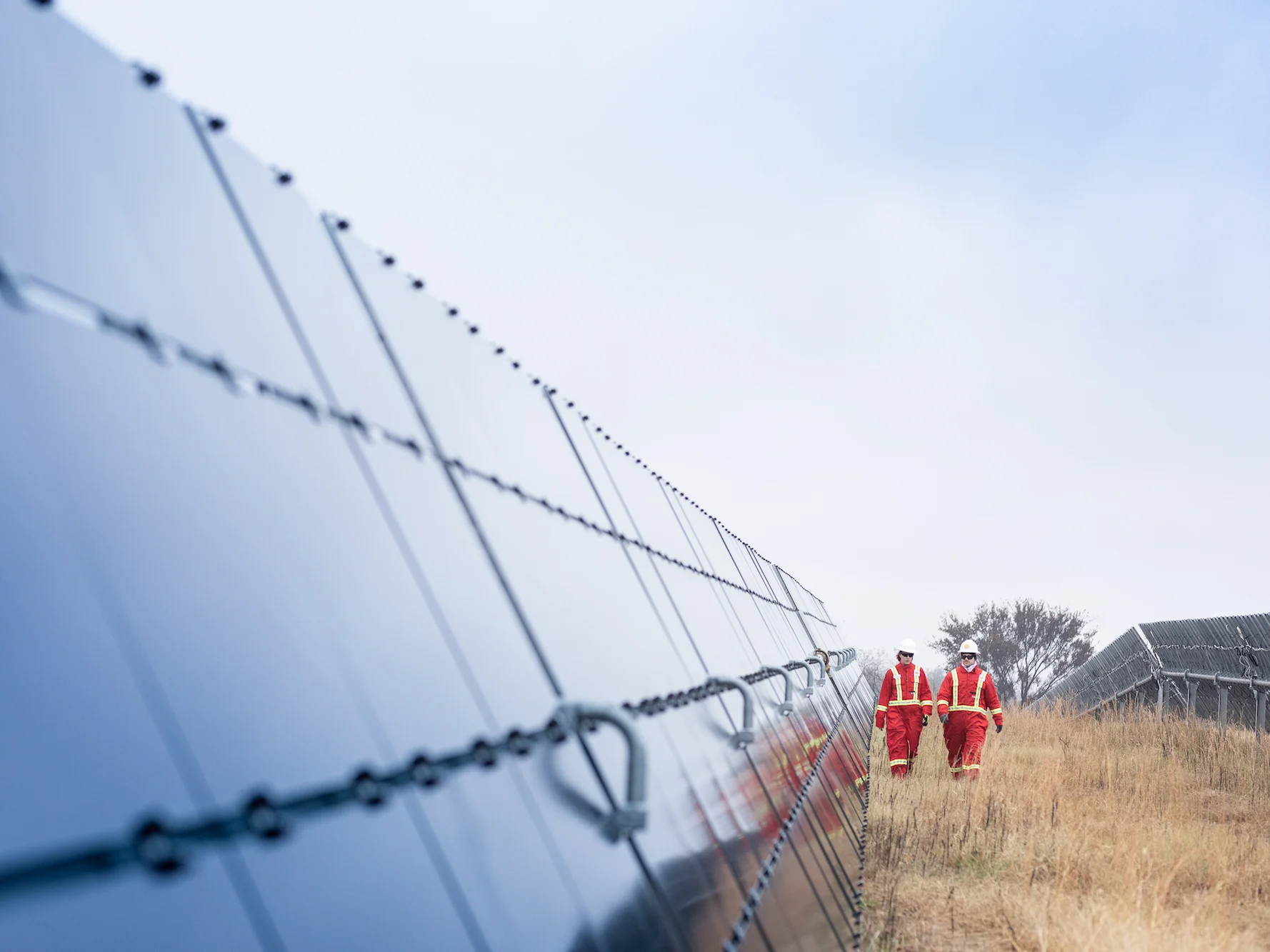 Two workers in red safety uniforms walking alongside rows of solar panels in an open field