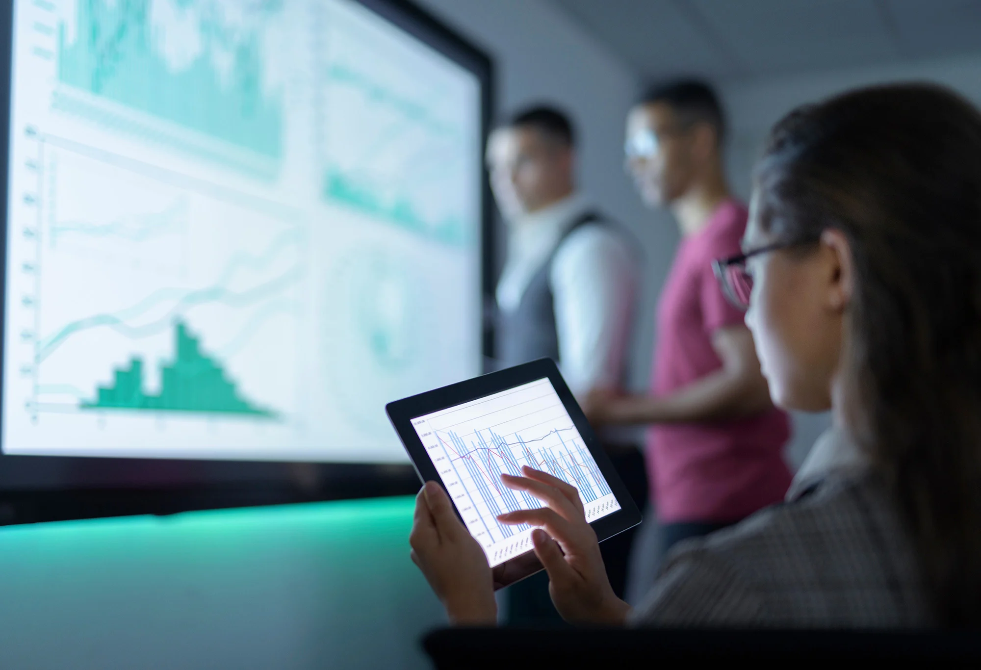 A person analyzing data on a tablet with graphs displayed on a large screen in the background, while two others are standing and discussing the data.