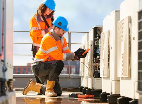 Two workers in safety gear inspecting and testing HVAC units on a rooftop.