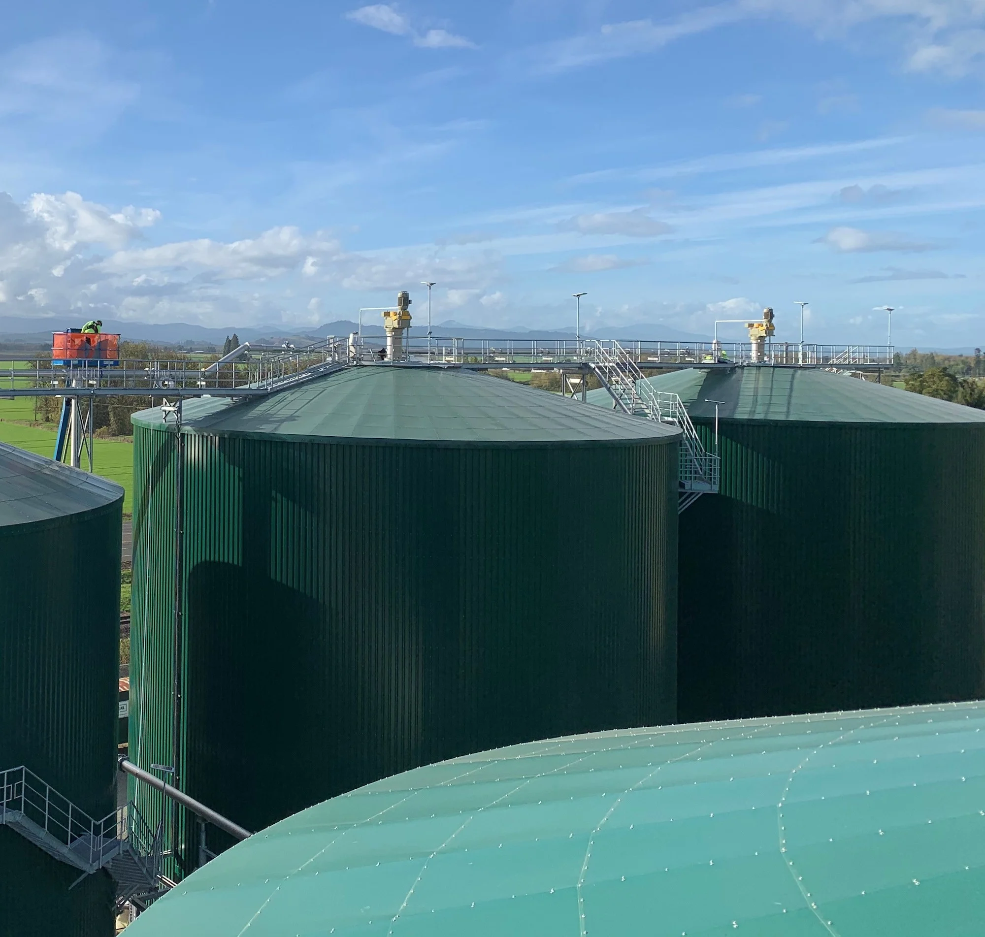Large green industrial storage tanks at a biogas or agricultural facility, set against a backdrop of fields and a partly cloudy sky.
