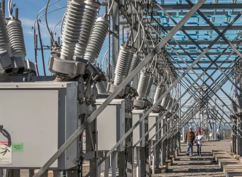 An outdoor electrical substation with rows of high-voltage equipment and two workers walking through the site