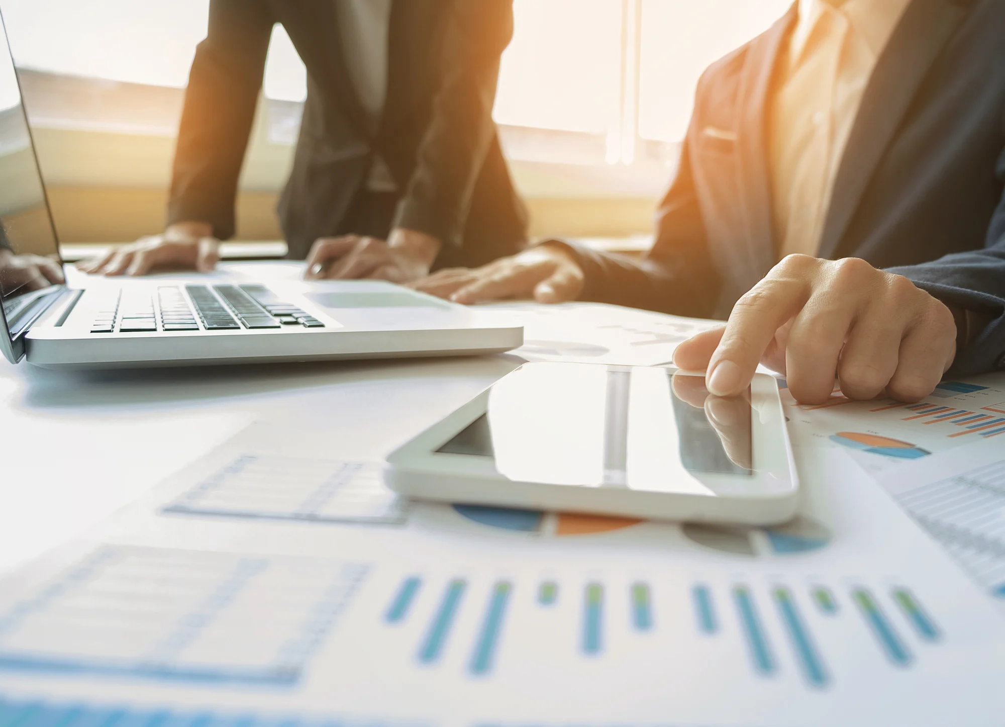 Close-up of business professionals working with a tablet and laptop, surrounded by charts and graphs on a desk