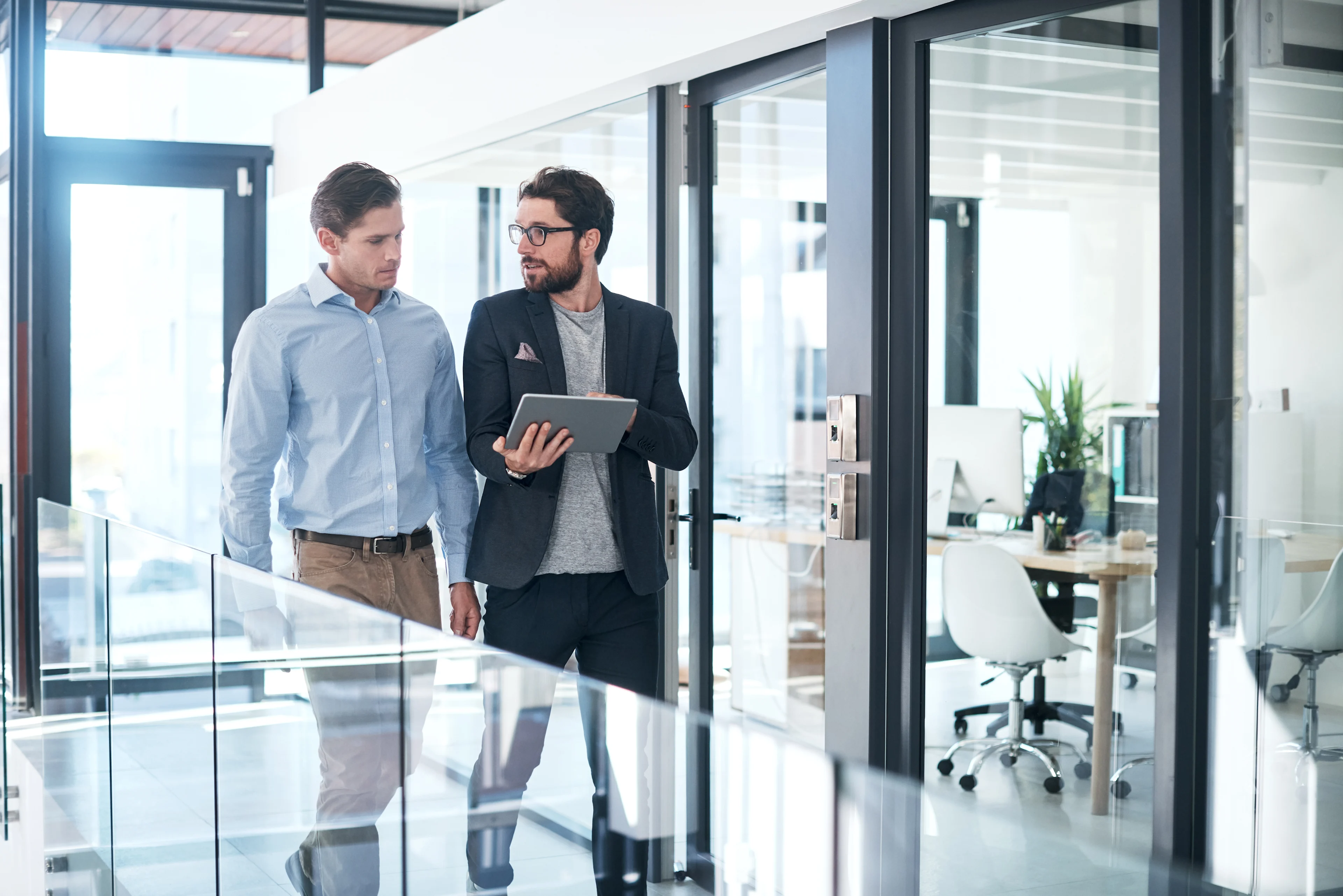 Two men are walking through an office hallway, engaged in a discussion while one of them holds a tablet.