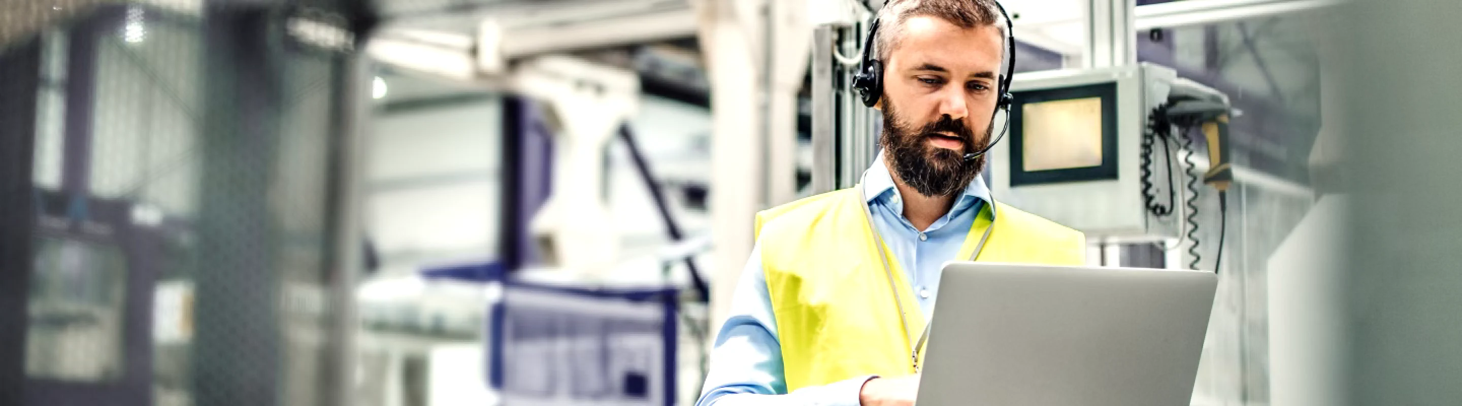 An engineer wearing a headset and safety vest working on a laptop in an industrial setting.