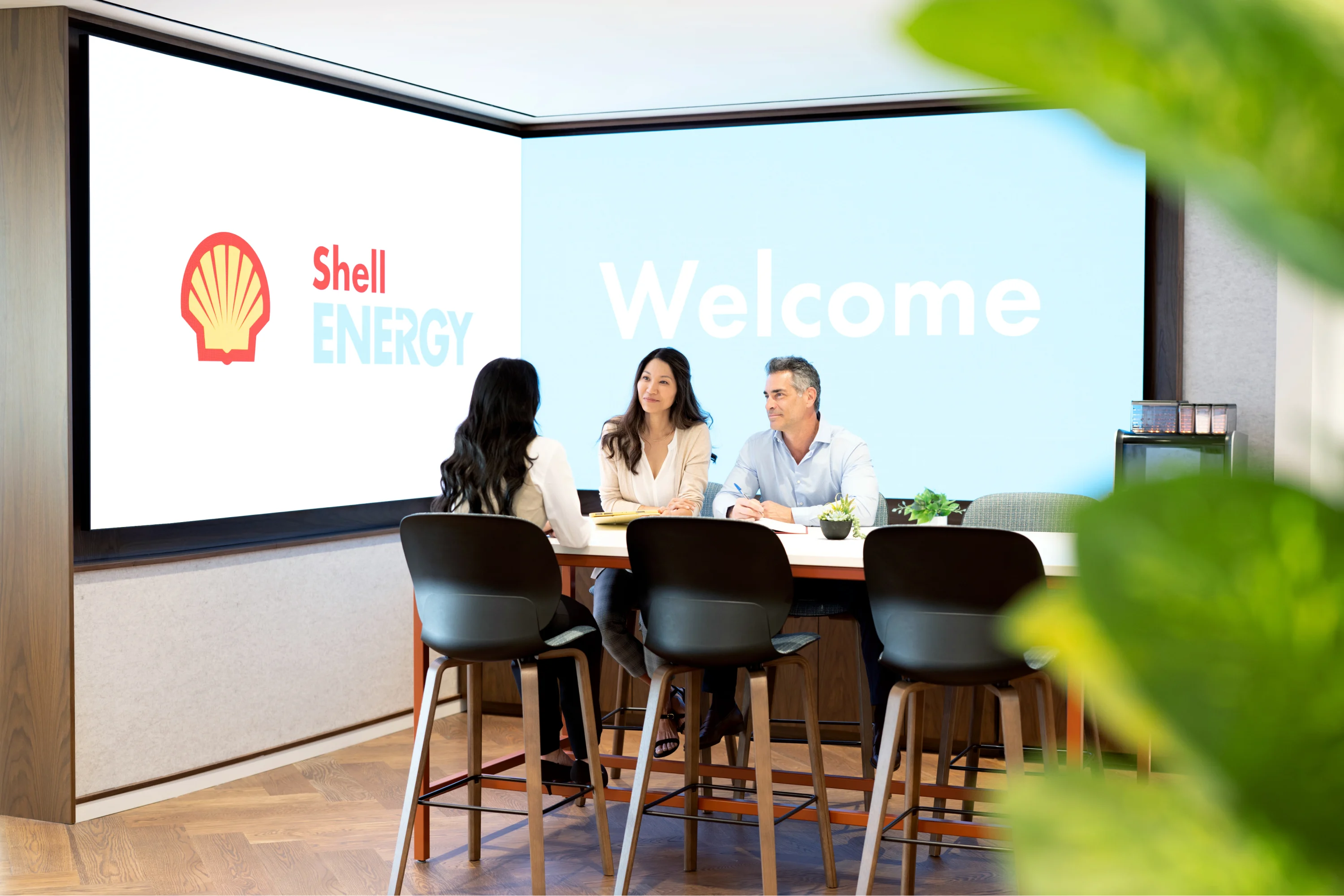 Three people having a meeting in a modern conference room with 'Shell Energy' and 'Welcome' displayed on large screens in the background