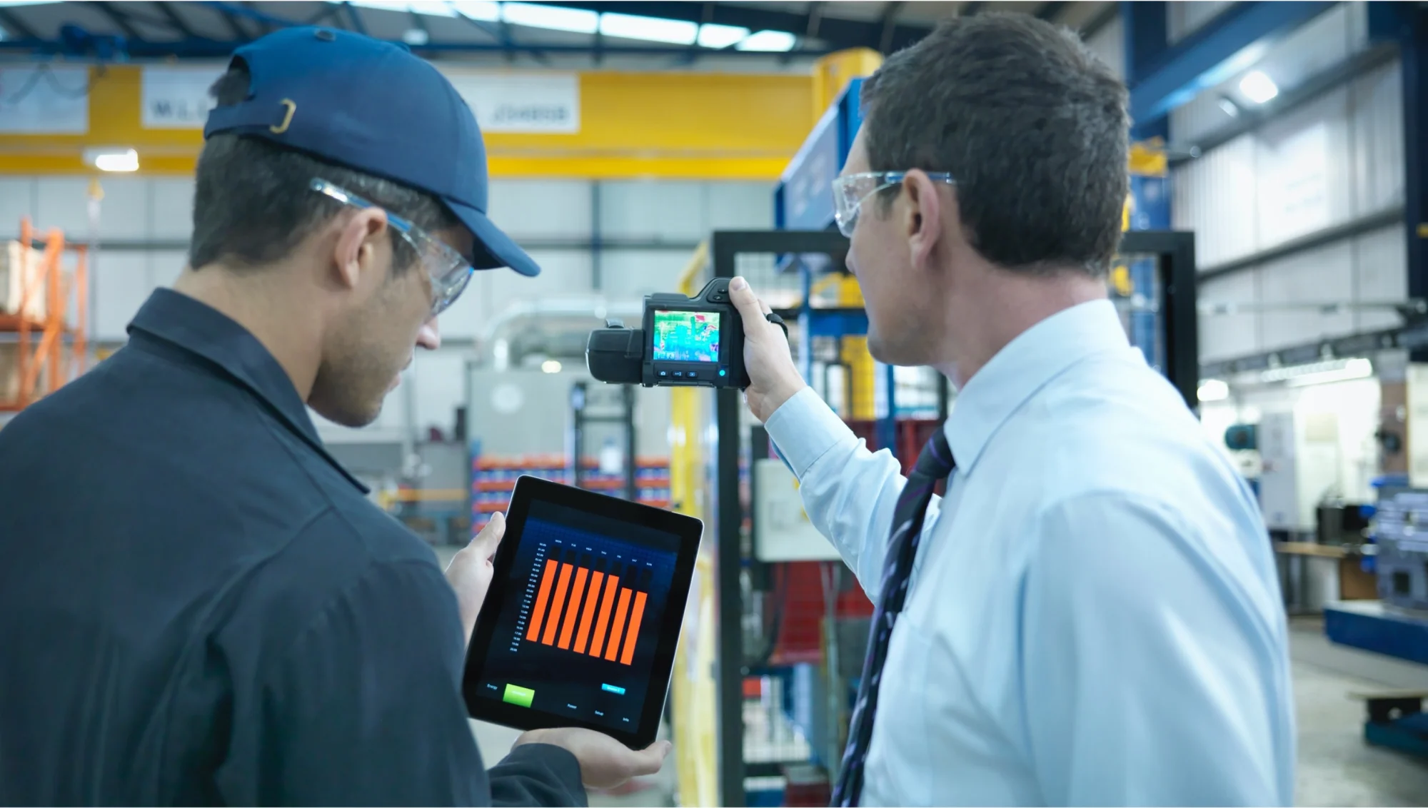 Two individuals in a factory, one holding a tablet with data and the other using a thermal imaging camera to inspect machinery.
