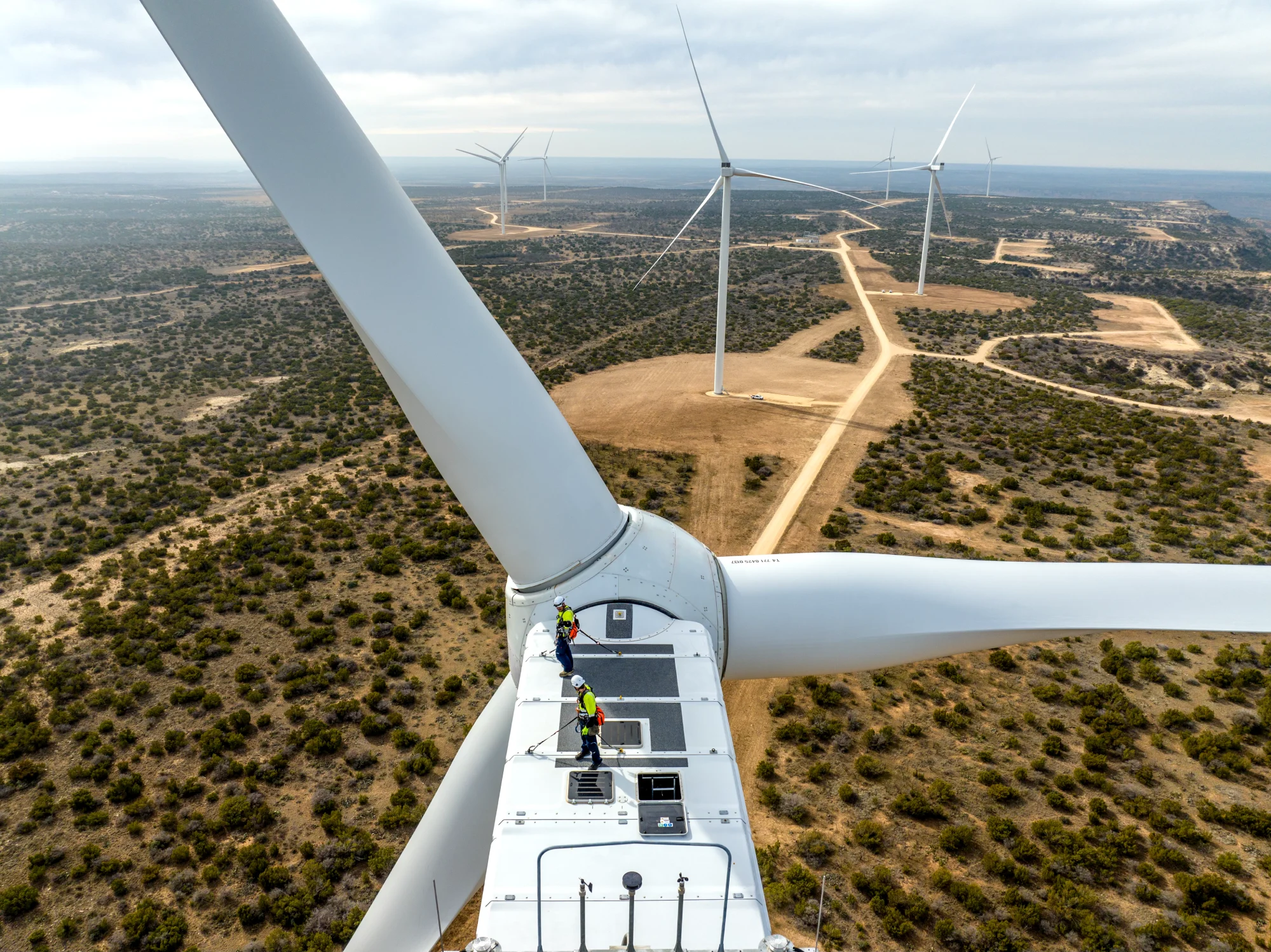 Two technicians wearing safety gear working on top of a wind turbine, surrounded by a landscape of multiple turbines